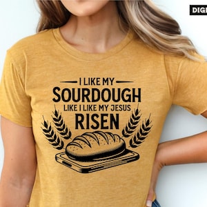 May include: A yellow t-shirt with a black graphic design that says "I like my sourdough like I like my Jesus risen". The graphic also includes a loaf of bread on a cutting board with wheat stalks on either side.