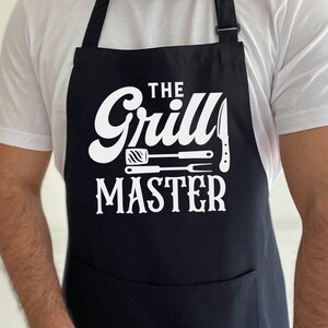 May include: Black apron with a white graphic that reads "THE Grill MASTER" with images of grilling tools. The apron has a front pocket and adjustable neck strap. The apron is worn over a white t-shirt.