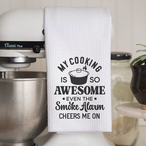May include: White kitchen towel with black text that reads "My cooking is so awesome even the smoke alarm cheers me on". The text is surrounded by a black circle with a small pot and flames inside.