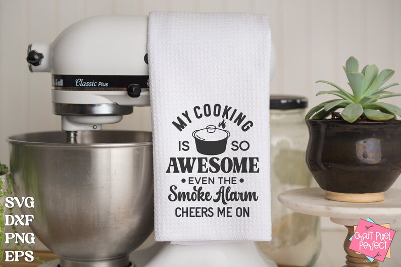 May include: White kitchen towel with black text that reads "My cooking is so awesome even the smoke alarm cheers me on". The text is surrounded by a black circle with a small pot and flames inside.