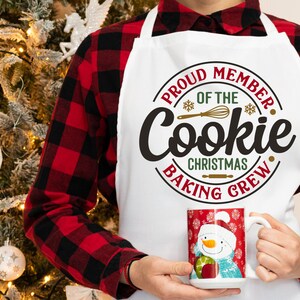 May include: A white apron with the text "Proud Member of the Cookie Christmas Baking Crew" in a red and gold circle. The apron is being worn by a person wearing a red and black plaid shirt.