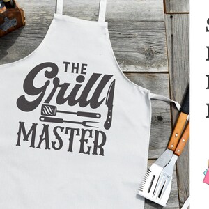May include: White apron with the text "The Grill Master" in a bold, dark gray font. The design includes illustrations of grilling tools. The apron is displayed on a rustic wooden surface, with grilling utensils and a wooden caddy in the background.