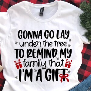 May include: White t-shirt with black text that reads "Gonna go lay under the tree to remind my family that I'm a gift". The text is surrounded by small red gift boxes and gray stars.