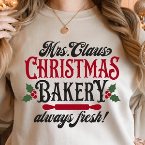 K&ouml;nnte beinhalten: Ein wei&szlig;er Pullover mit dem Text "Mrs. Claus Christmas Bakery always fresh!" in schwarzer und roter Schrift. Der Text ist von gr&uuml;nen Stechpalmenbl&auml;ttern und roten Beeren umgeben. Unter dem Text befindet sich ein roter Nudelholz.