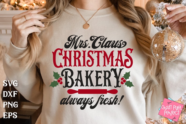 K&ouml;nnte beinhalten: Ein wei&szlig;er Pullover mit dem Text "Mrs. Claus Christmas Bakery always fresh!" in schwarzer und roter Schrift. Der Text ist von gr&uuml;nen Stechpalmenbl&auml;ttern und roten Beeren umgeben. Unter dem Text befindet sich ein roter Nudelholz.