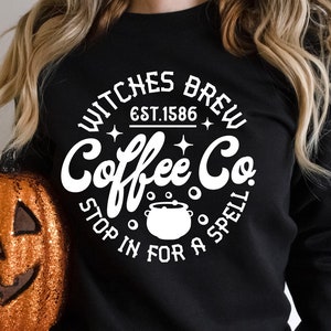 以下が含まれることがあります： 「Witches Brew Coffee Co. Est. 1586 Stop in for a Spell」と書いてある白い円形のデザインが施された黒いスウェットシャツ。釜のグラフィックが描かれている。