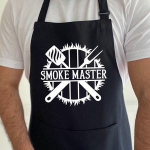 May include: A black apron with a white design of crossed grilling tools and a circle with flames around it. The text "SMOKE MASTER" is inside the circle.
