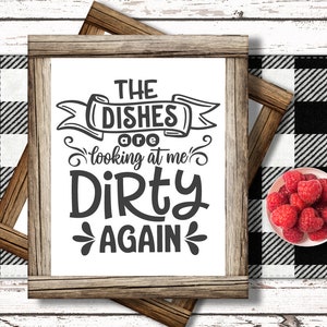 May include: A framed sign with the text "THE DISHES are looking at me DIRTY AGAIN" in black lettering. The sign is set against a black and white checkered background, with a small bowl of raspberries on the right.