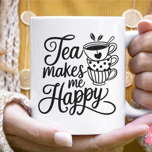 May include: White ceramic mug with a black graphic that says "Tea makes me happy" and a stack of teacups with a tea bag and spoon.