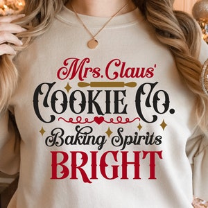 May include: A white sweatshirt with the text "Mrs. Claus' Cookie Co. Baking Spirits Bright" in red, black, and gold lettering. The text is in a festive, handwritten font.