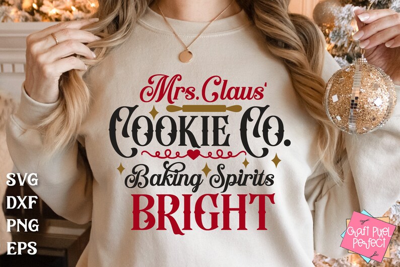 May include: A white sweatshirt with the text "Mrs. Claus' Cookie Co. Baking Spirits Bright" in red, black, and gold lettering. The text is in a festive, handwritten font.