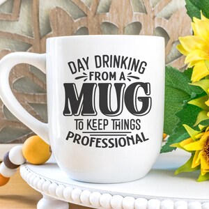 May include: White ceramic mug with black text that reads "Day Drinking From A Mug To Keep Things Professional". The mug is sitting on a white pedestal with a beaded garland and sunflowers in the background.