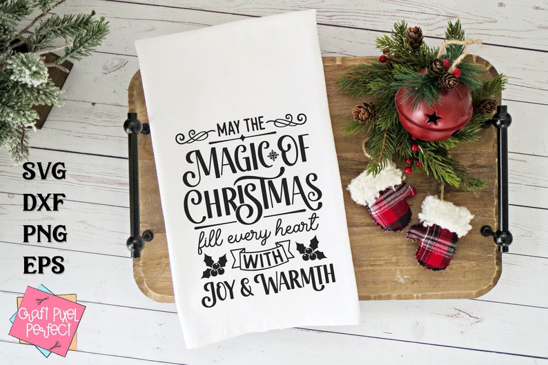 May include: A white kitchen towel with black text that reads "May the magic of Christmas fill every heart with joy & warmth". The towel is displayed on a wooden tray with a red bell ornament and red and white mittens.