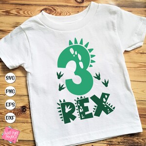 Op de afbeelding: Wit T-shirt met een groen dinosaurusontwerp. Het ontwerp bevat het nummer 3, dinosaurusvoetstappen en het woord "REX". Dit T-shirt is waarschijnlijk voor de derde verjaardag van een kind.