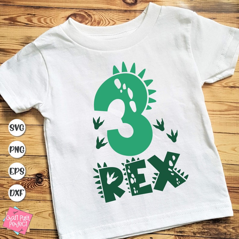 Op de afbeelding: Wit T-shirt met een groen dinosaurusontwerp. Het ontwerp bevat het nummer 3, dinosaurusvoetstappen en het woord "REX". Dit T-shirt is waarschijnlijk voor de derde verjaardag van een kind.