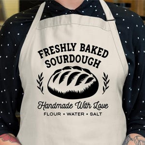 May include: A cream apron featuring the words "FRESHLY BAKED SOURDOUGH" in black, a bread illustration, and the phrase "Handmade With Love". The apron also includes the text "FLOUR &bull; WATER &bull; SALT". The apron is a digital design.