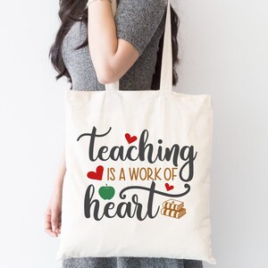 May include: A white tote bag with the text "teaching is a work of heart" in black. The text is surrounded by red hearts and a green apple. There are also two stacks of books in the design.