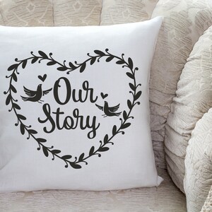May include: White throw pillow with black text that reads "Our Story" inside a heart-shaped wreath with two birds.