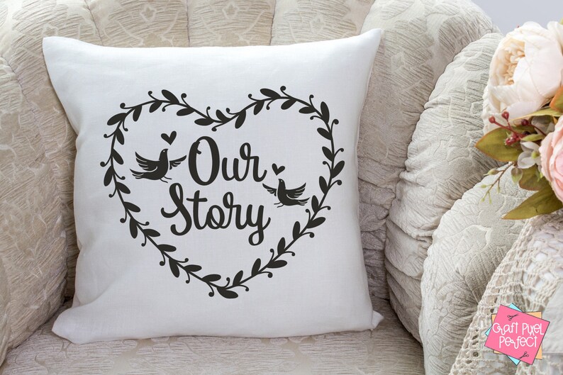 May include: White throw pillow with black text that reads "Our Story" inside a heart-shaped wreath with two birds.