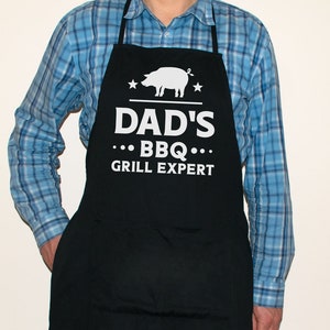 May include: A black apron with white text that reads "Dad's BBQ Grill Expert" and a white silhouette of a pig. There are two white stars on either side of the pig.