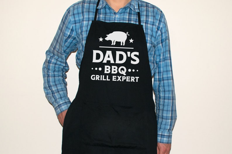 May include: A black apron with white text that reads "Dad's BBQ Grill Expert" and a white silhouette of a pig. There are two white stars on either side of the pig.