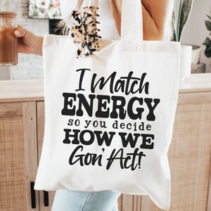 May include: A white tote bag with the text "I Match ENERGY so you decide HOW WE Gon' Act!" in black. The bag is held in front of a wooden cabinet. A glass with a drink is held in the other hand.
