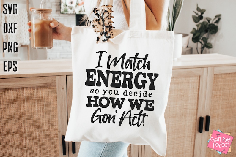 May include: A white tote bag with the text "I Match ENERGY so you decide HOW WE Gon' Act!" in black. The bag is held in front of a wooden cabinet. A glass with a drink is held in the other hand.