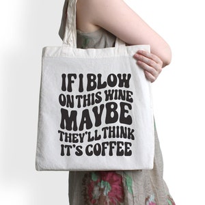 Op de afbeelding: Een witte tote bag met zwarte tekst die "Als ik op deze wijn blaas, denken ze misschien dat het koffie is" zegt.