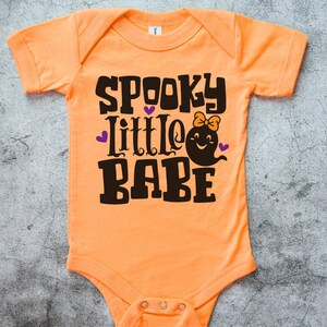 May include: Orange baby onesie with the text "Spooky Little Babe" in black with a ghost graphic and purple hearts.
