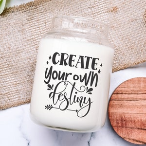 May include: White candle jar with a black text overlay that reads "CREATE your own destiny" with an arrow pointing to the right. The candle jar is sitting on a white surface with a brown wooden coaster.