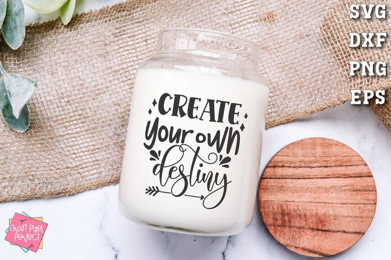 May include: White candle jar with a black text overlay that reads "CREATE your own destiny" with an arrow pointing to the right. The candle jar is sitting on a white surface with a brown wooden coaster.