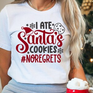 May include: A white t-shirt with the text "I ate Santa's cookies #noregrets" in red and gray lettering. A red Santa hat and a chocolate chip cookie are also included in the design.