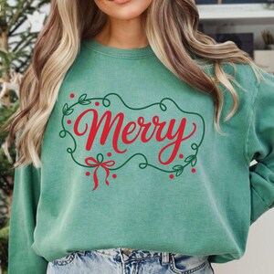 May include: A teal green sweatshirt with the word "Merry" in red script, framed by a green leafy design with red berries and a red bow. The words "DIGITAL DESIGN" are in the top left corner.