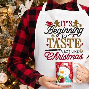 May include: White apron with red and gold text that reads "It's Beginning to Taste A Lot Like Christmas". The apron has a gingerbread man design and is worn by a person wearing a red and black plaid shirt.