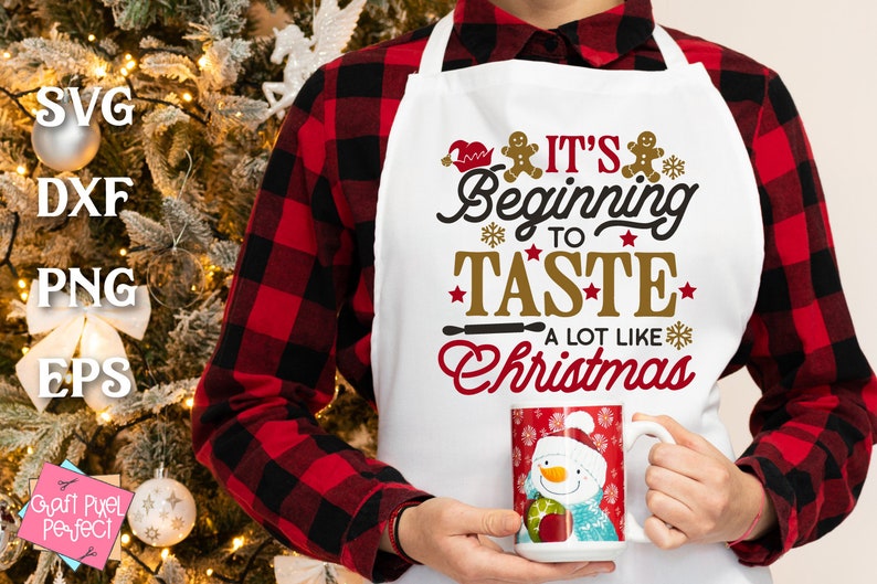 May include: White apron with red and gold text that reads "It's Beginning to Taste A Lot Like Christmas". The apron has a gingerbread man design and is worn by a person wearing a red and black plaid shirt.