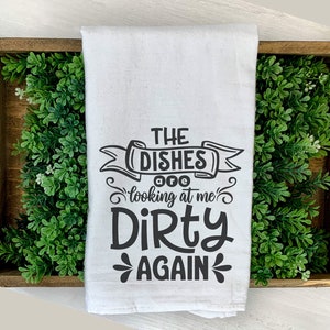 May include: White kitchen towel with the text "THE DISHES are looking at me DIRTY AGAIN" in black. The towel is displayed in a wooden tray with green foliage.