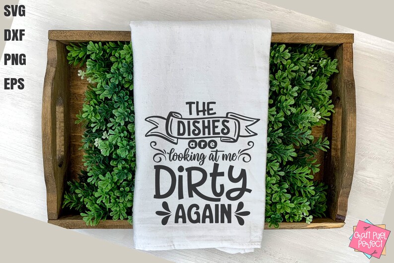 May include: White kitchen towel with the text "THE DISHES are looking at me DIRTY AGAIN" in black. The towel is displayed in a wooden tray with green foliage.