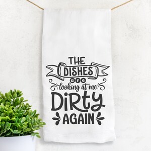 May include: White kitchen towel with the text "THE DISHES are looking at me DIRTY AGAIN" in black. The towel is hanging from a string. A small green plant in a white pot is in the lower left corner.