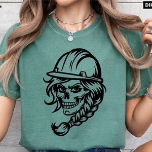 Op de afbeelding: Een zwart-wit grafisch ontwerp van een schedel met een helm op. De schedel heeft lang haar dat is gevlochten. Het ontwerp is op een groen T-shirt.