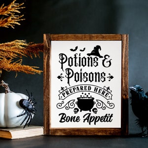 Pu&ograve; includere: Un cartello di legno con una grafica in bianco e nero di un calderone, pipistrelli e un cappello da strega. Il testo recita "Pozioni & Veleni Preparati Qui. Buon appetito".