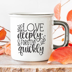 May include: White ceramic mug with a black handle. The mug has black text that reads "Love deeply: & forgive quickly". The mug is sitting on a wooden surface with autumn leaves around it.
