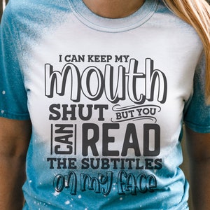 K&ouml;nnte beinhalten: Wei&szlig;es und blaues T-Shirt mit Batikmuster und dem Text "I can keep my mouth shut but you can read the subtitles on my face" in Schwarz. Das Shirt hat auch die Texte SVG, DXF, PNG und EPS.