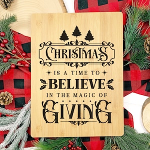 May include: A wooden cutting board with the text "Christmas is a time to believe in the magic of giving" in black lettering. The board is surrounded by red and black plaid fabric, pine branches, and pine cones.