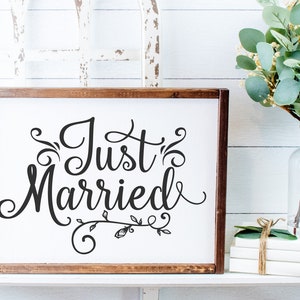 May include: A white wooden sign with a brown frame, featuring the text "Just Married" in a decorative script font with floral embellishments.