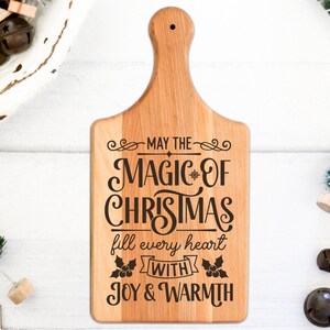 Puede incluir: Una tabla de cortar de madera con un dise&ntilde;o navide&ntilde;o. El texto dice "Que la magia de la Navidad llene cada coraz&oacute;n de alegr&iacute;a y calidez".
