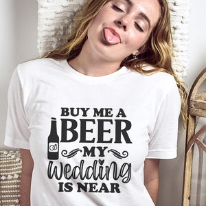 May include: White t-shirt with black text that reads "Buy me a beer my wedding is near". The text is stylized with a beer bottle graphic and wedding rings.