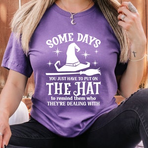 May include: Purple t-shirt with a white graphic of a witch's hat and the text "Some days you just have to put on the hat to remind them who they're dealing with."