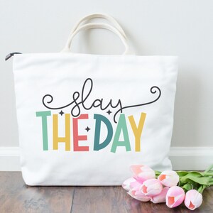 May include: A white tote bag with the text "slay the day" in colorful letters. The letters are outlined in black and have a retro, 70s style. The bag is sitting on a wooden surface with a bouquet of pink tulips.