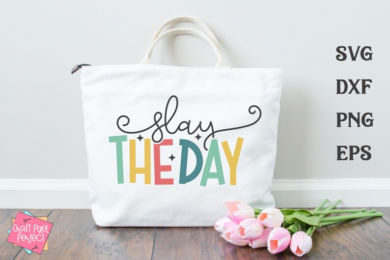 May include: A white tote bag with the text "slay the day" in colorful letters. The letters are outlined in black and have a retro, 70s style. The bag is sitting on a wooden surface with a bouquet of pink tulips.