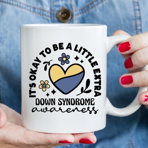 May include: White ceramic mug with a black and blue design. The design features a heart with yellow and blue colors, flowers, and the text "It's okay to be a little extra Down Syndrome awareness".